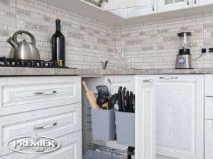 Modern kitchen with white cabinetry, featuring pull-out cutlery cubbies, a sleek backsplash, and a Premier Kitchen & Bath logo in the corner